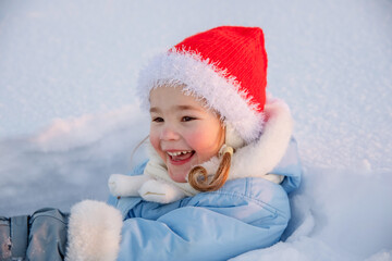 A joyful child girl sits in a snowdrift in a blue jacket and a red cap.
