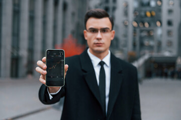 Showing phone with crypto graphs in it. Businessman in black suit and tie is outdoors in the city