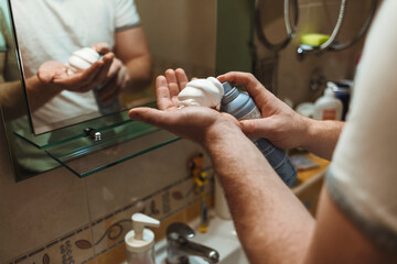 Young man looking in the mirror and preparing skin for shaving. Man getting ready for shaving in front of the mirror in his bathroom. Happy young man looking at mirror and applying shaving cream