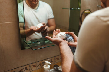 Close-up shot of handsome man standing at the bathroom mirror and applying shaving foam while preparing shaving. Male applying shaving cream to his face in the bathroom at home.