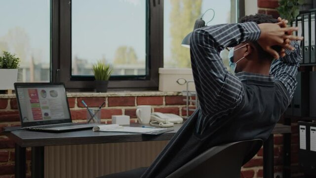 Business man taking break from work project in office, wearing face mask. Employee relaxing at desk, on timeout after working with laptop to plan executive career during pandemic.