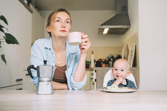 Working mother with little baby. Young tired woman with coffee and baby having breakfast in kitchen. Modern freelancer mom and her child after sleepless night. Work-life or work-family balance