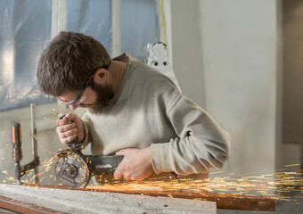 a man with a beard in glasses cuts a metal profile indoors with an angle grinder, sparks fly.