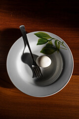 Still life of minimal food with boiled egg in a white plate with fork on a wooden table in the sunlight.