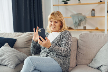 Sitting and holding phone. Senior woman with blonde hair is at home