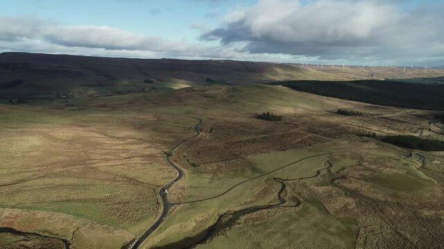 Aerial footage of a white van winding road through moorland with wind turbines turning on the summit of a 