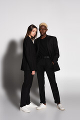 full length view of blonde african american man and brunette woman in black suits on grey with shadow.