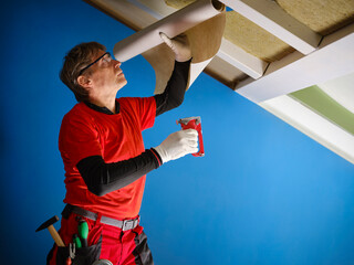 Craftsman in work clothes on a ladder, applies the anti-vapor sheet with a stapler to cover the...