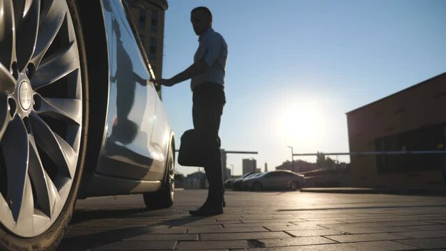 Young confident businessman in formal wear going to modern electric car. Successful entrepreneur with briefcase opening door and getting inside to new electrical SUV vehicle. Sunlight at background