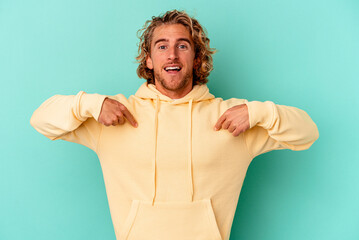 Young caucasian man isolated on blue background surprised pointing with finger, smiling broadly. - obrazy, fototapety, plakaty