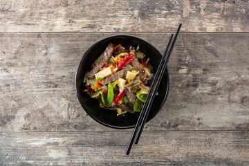 Beef, vegetables and sesame seeds in black bowl on wooden table. Top view. Copy space