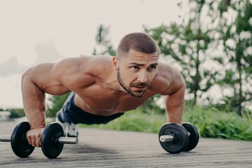 Fototapeta na wymiar Young motivated sportsman stands in plank pose, trains muscles with barbells, focused into distance with determined expression, has naked torso, has strong muscular body. Athlete bodybuilder outdoor