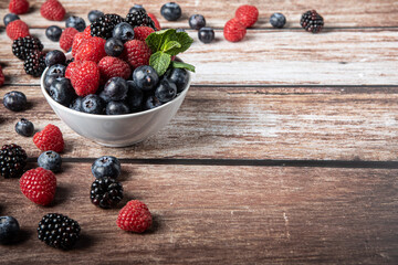 Bowl of red berries on a wooden background with copy space on the right. Freshly picked organic blackberries, blueberries and raspberries. Healthy food and diet.