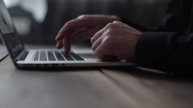 Close-up hands of unrecognizable mature male typing on laptop keyboard at table in dark room. Closeup of aged business man working on computer indoors, browsing Internet. Tracking shot in slow motion.