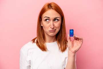 Young caucasian woman holding a batteries isolated on pink background confused, feels doubtful and unsure.