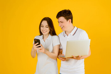 Portrait of a young happy couple man and woman in white t-shirts using smartphones and notebook on a yellow background.