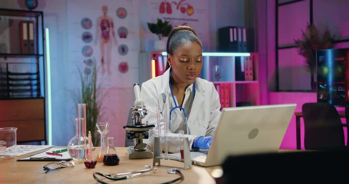 Attractive confident skilled african amrican female chemist sitting at her workplace in lab with different test tubes with solutions and entering datas of research into laptop