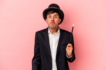 Young caucasian wizard man holding a wand isolated on pink background shrugs shoulders and open...
