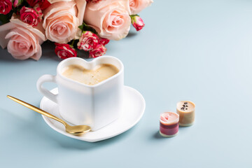 Cup of coffee in shape of heart with chocolate candies and bouquet of roses on blue background. Love concept.