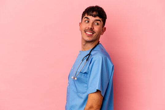 Young mixed race nurse man isolated on pink background looks aside smiling, cheerful and pleasant.