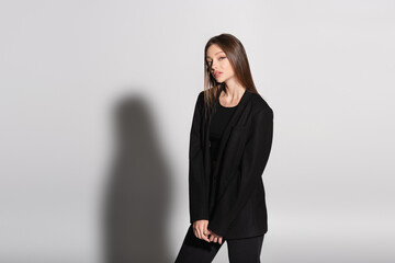 stylish woman in black clothes looking at camera while standing on grey with shadow.