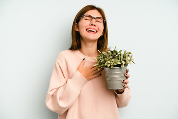 Young English woman holding a plant isolated on blue background laughs out loudly keeping hand on chest.