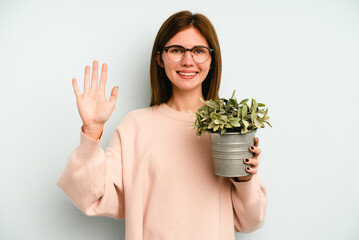 Young English woman holding a plant isolated on blue background smiling cheerful showing number five with fingers.