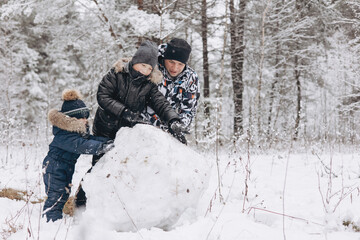 Fototapeta na wymiar Happy father and sons rolling big snowball together for snowman in snowy winter forest. Man Joyful teenager and little boy playing and having fun on family walking. Wintertime activity outdoors.