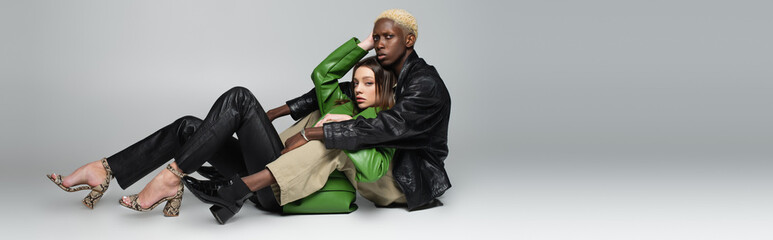 stylish brunette woman and blonde african american man looking at camera while sitting on grey,...
