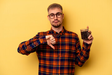 Young caucasian man holding coffee jar isolated on yellow background showing a dislike gesture,...