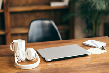 Workplace concept. Silver laptop with cup, headphones and notebook on a wood table in a home interior. Freelance home office.