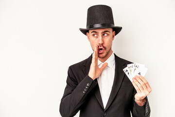 Young caucasian wizard man holding a magic cards isolated on white background is saying a secret hot braking news and looking aside