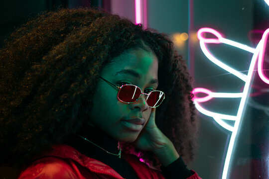 Portrait of happy beautiful girl with sunglasses sitting in her longboard and smiling at the neon light. Urban scene, city life