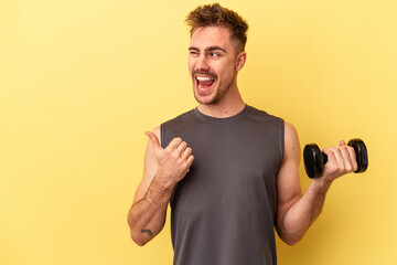 Young sport man holding a dumbbell isolated on yellow background points with thumb finger away, laughing and carefree.