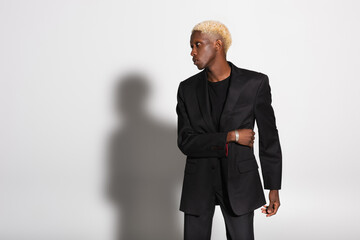 blonde african american man in black suit and silver bracelet looking away on grey with shadow.