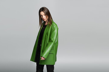 young and stylish woman in green leather jacket looking at camera isolated on grey.