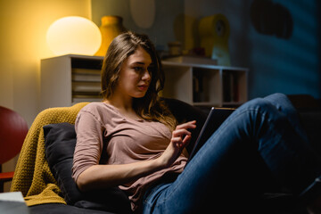 young woman relaxing on sofa at home. she is using digital tablet. night evening scene