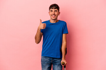 Young mixed race man with crutches isolated on pink background smiling and raising thumb up