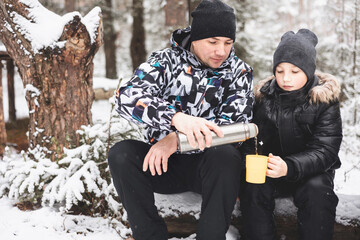 Father and sons drinking tea from thermos and talking sitting together on log in winter snowy forest. Happy man and teenage boys having picnic in winter season outdoors. Local travel. Slow life