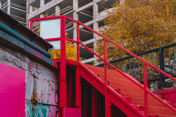 Red wooden staircase on the street with construction site on the background. Urban. City. Development. Project. Painted. Tree. Autumn