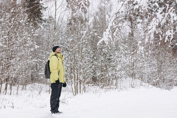 Fototapeta na wymiar Rear view of young man with backpack in yellow jacket walking in snowy pine forest in winter. People from behind. Local travel, exploring nature.
