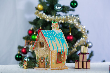 Gingerbread house with blue turquoise icing roof on a table next to miniature gift boxes and a decorated Christmas tree in the background