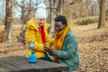 cheerful interracial couple looking at cellphone outdoors. He shows her something interesting, they spend beautiful moments in the autumn forest. They are dressed warmly