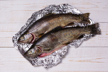 Raw trout fish on aluminum foil on wooden background, top view, flat lay image.