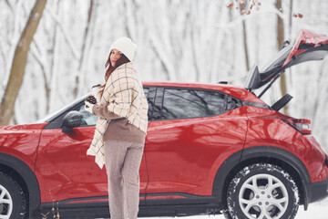With cup of drink. Beautiful young woman is outdoors near her red automobile at winter time