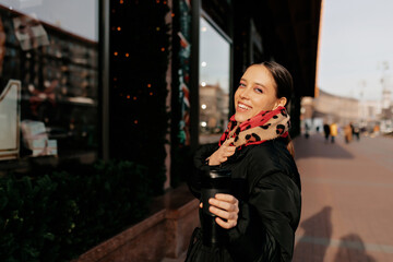 Close-up of cute european woman looking at camera on blurred city street background. Happy smiling girl covering turn around at camera with coffee while resting in the city