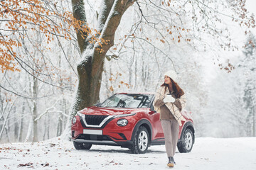 Rural scene. Beautiful young woman is outdoors near her red automobile at winter time