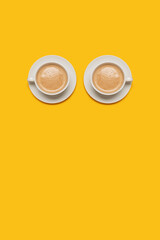 Two cups of coffee with foam on top in white mugs on white saucers on a yellow background.  Room for text and copy space in a vertical image with a top composition
