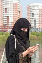 A Muslim woman in national clothes with her face covered with a phone in her hand against background large city.