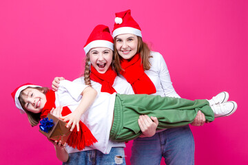 Seasonal Holidays Ideas. Three Happy Caucasian Girls Wearing Santa Hats Having Fun While Having Teenare Girl Lifted On Hands With Wrapped Golden Gift.